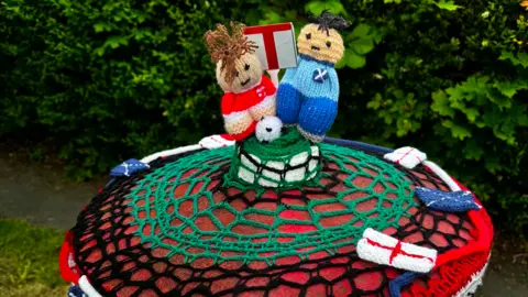 Robyn Cook Knitted footballers in England and Scottish team colours line up on top of a crocheted cover on a red post box