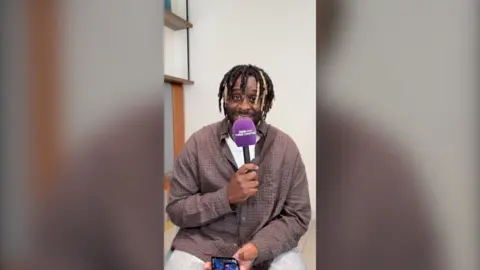 Myles Smith is smiling, staring at the camera. He is hold a BBC Three Counties Radio branded purple mic. He is wearing a checkered brown shirt. 