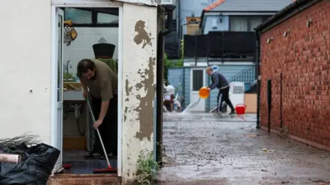 A man in the foreground is sweeping water out of his property. In the background is a man bailing out water with an orange bucket