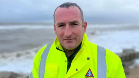 Dan Hurd in a high visibility jacket stood on the beach with the sea in the background. Dan has short dark hair and stubble on his face.