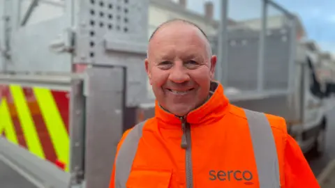 Harry Archer pictured smiling and stood in front of a silver van. He is bald and wears an orange hi-vis serco jacket.