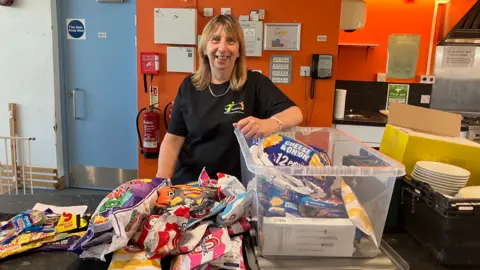 Julie Nerton smiling at the camera inside the Rollercoaster youth club standing behind a pile of crisps on a kitchen worktop