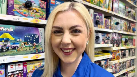 A head and shoulders shot of employee Jessica Payne looking straight at the camera. She is wearing the royal blue shirt which is the uniform of The entertainer. She has long blonde hair and is smiling. Behind her are shelves of boxes of toys. 