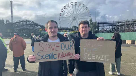Cash Murphy/BBC Protest organiser Nick Chamberlain and protester Daniel Bishop pictured in front of the Scenic Railway in Dreamland, Margate. They are each holding signs. Nick's sign says: "Save our Scenic Railway" and Daniel's sign says: "Save the Scenic."