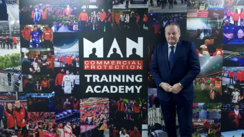 Man in a blue suit, white shirt, blue tie standing next to a wall showing images of security staff at work. In the centre of the images is a logo reading MAN Commercial Protection training academy