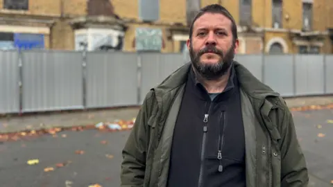 A man with brown hair and a beard and wearing a blue fleece and green coat is standing in front of boarded up terraced houses. The properties have a metal fence around them. 