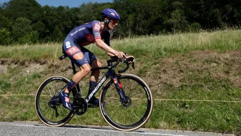 Reuters Oscar Onley on a Lapierre bike with a blue cycle kit on, blue helmet and shows. There is a grass banking behind him.