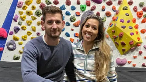 A couple wearing athletic clothing sit on a mat in front of an indoor bouldering wall