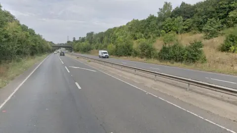 Google A602 where it changes from a dual carriageway to a single lane carriageway, with a white arrow marking guiding drivers into the single lane. There is a central reservation in the middle. There are banks either side with trees and a bridge over the road in the distance. A few cars are seen in the distance.