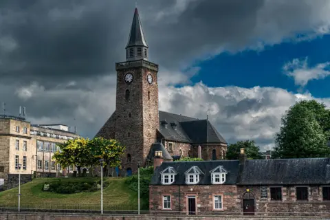 Getty Images A view of Inverness' Old High Church from across the River Ness. The church is a stone-built kirk with a steeple with clock faces near the top. There is a row of traditional cottages on the street below.