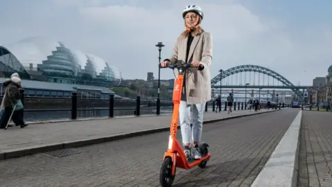 Neuron Mobility A woman with long blonde hair is riding an orange Neuron scooter along Newcastle's Quayside. She is wearing jeans, a beige coat and a white helmet. The Tyne Bridge and the Glass House in Gateshead can be seen across the river.