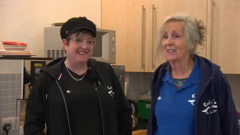 Katie and Sue Robinson pictured in the kitchen area of a church hall, dressed in branded t-shirts and hooded jumpers of their group 