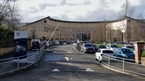 Google Sand coloured building at Bangor Academy in the background, with various cars in front and white and blue signage to the left. There are railings on both sides.