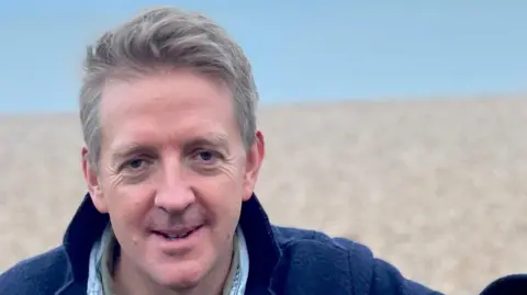 Supplied A close-up head and shoulders image of Dr John Paul-Stonard standing on a beach. He is wearing a navy blue jacket and smiling while looking into the camera.