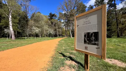 Shaun Whitmore/BBC A wooden sign is on the right and it has a black and white picture and text. There is a path on the left which leads to a woodland area. 
