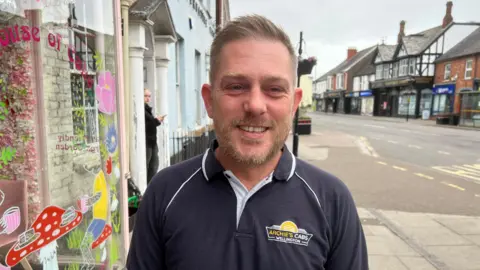 Paul Higginbottom is outside House of Cake in Wellington High Street. He is wearing an "Archie's Cabs" T-shirt.