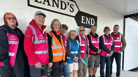 Yvonne Turner Yvonne Turner stood in the middle of a group of Parkrun volunteers who are wearing pink vests. One woman is wearing an orange hi-vis and Yvonne is wearing a blue hi-vis.