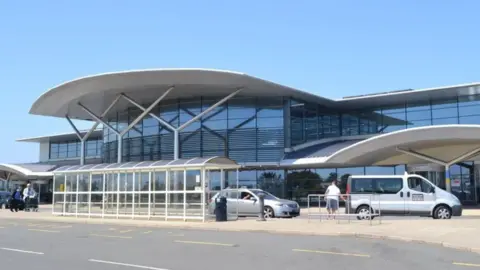 BBC Guernsey airport - a building made predominantly out of glass windows, a taxi rank in front with two taxis, a road in front of it on a sunny day