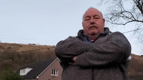 A bald man with a moustache wearing a grey fleece, looking angry with his arms crossed across his body. Behind him are houses and hills.