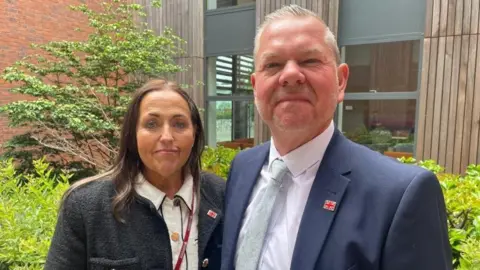 A woman wearing a white top and grey jacket, stood alongside a man wearing a blue suit, white shirt and green tie. Behind them is a large council building.