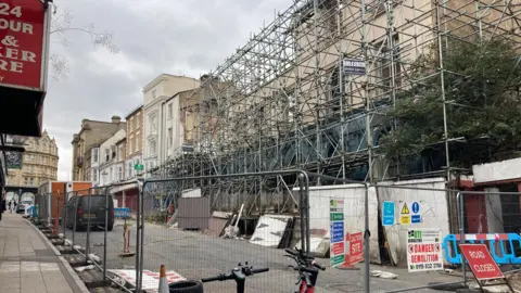 BBC A street with silver barriers blocking the entrance to the road, with a red sign that reads "Road Closed". On the right is a large amount of erected scaffolding across a partly-demolished building. 