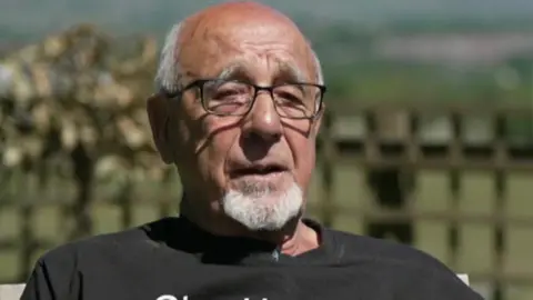 Head and shoulders image of John Morris sitting in the garden in the sunshine in a black tshirt. He is wearing glasses and has short white receeding hair and a goatee