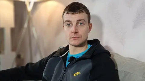 Philip Mitchell, a man with short, dark hair, looks to the camera in a sitting room. He is wearing a bright blue t-shirt under a black and grey sports jacket. He is sitting on a grey sofa and a standard lamp is partially visible behind his head.