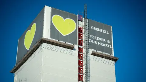 The top section of Grenfell Tower, covered in white tarpaulin, with scaffolding along one side.