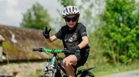 Red On Sports A young boy on a bike holds up two fingers for victory while pedalling a green bike. He is in a black top and shorts with a white helmet on and dark glasses.