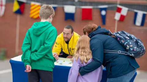 RNLI/Mike Milner A boy dressed in a green hoodie, with his back to the camera, speaks to a lifeboat station worker who is wearing a yellow jacket and sitting behind a blue desk. To the right, a woman wearing a blue coat and a rucksack has her arm around a small girl wearing a violet hoodie. In the background is a red brick wall with a line of colourful flags hanging from it.
