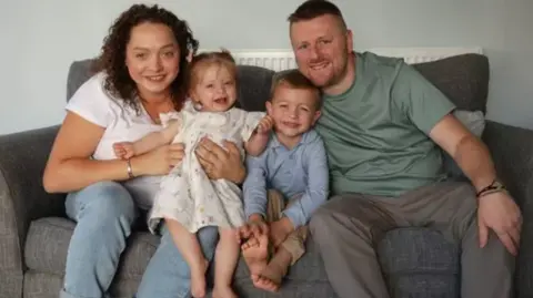Family handout The family sit on a grey couch. Colette has shoulder-length curly brown hair. She wears a white T-shirt and pale blue jeans. She is holding Hollie on her knee, she has short blonde hair and wears a white dress. Ted sits between Hollie and Kevin. He wears a blue long-sleeved polo shirt and beige trousers. He has short blonde hair. Kevin sits next to Ted, he wears a green T-shirt and grey trousers. They are all smiling at the camera.