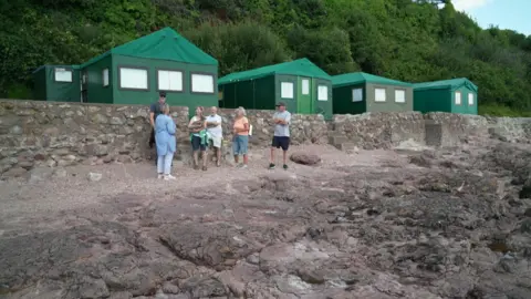 A group of people stand on a rocky beach. Behind them are four temporary green huts which are put there every summer.