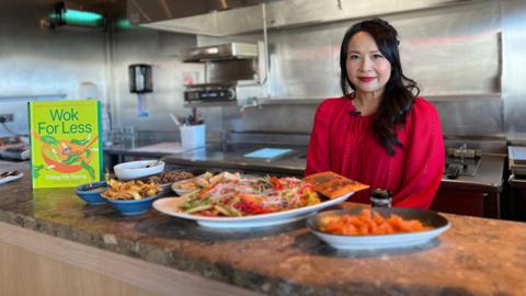 Ching-He Huang smiles at the camera with Chinese on a counter in front of her with her book to the left of the food