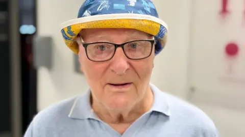 Michael Lyall with white hair wearing a Leeds Rhinos blue bucket hat and pale blue collared top. He is smiling.