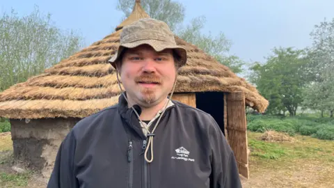 Arthur Randall looking at the camera. He is wearing a sunhat and a Flag Fen Archaeology Park-branded jumper. Behind him is the replica roundhouse, with layers of thatched roof over a circular wall. There is an open wooden door at the front. 