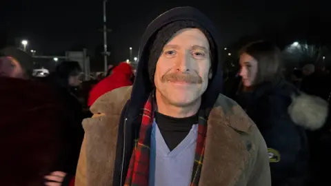 A man with a moustache wears a hat, hoodie and red tartan scarf, with a sheepskin coat on top, smiles at the camera.