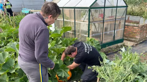 Two men pick vegetables from an allotment. One is standing by a vegetable patch wearing a grey top and jogging bottoms, while another, wearing a black t-shirt and black trousers, is crouched down tending to a large pumpkin. 