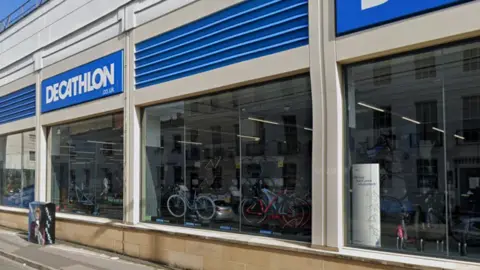 The side exterior of Decathlon, a sporting goods shop. There is blue and white branding above large windows, through which you can see lots of bikes arranged for sale.