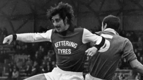 Kettering Town FC A black and white image of a Kettering Town footballer in action. He is mid-air competing for the ball, which is out of shot. An opposition player is behind him, as is the crowd.