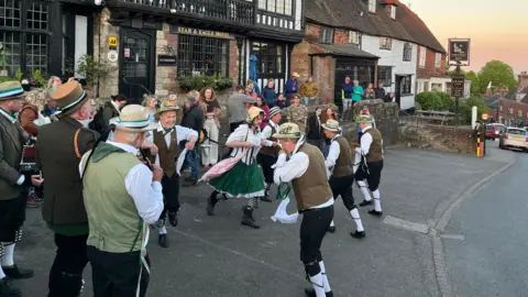 Several people in traditional Morris dancing costume, playing musical instruments and dancing in two lines. A small crowd stood outside a pub watches on, with an orange sunrise in the sky.