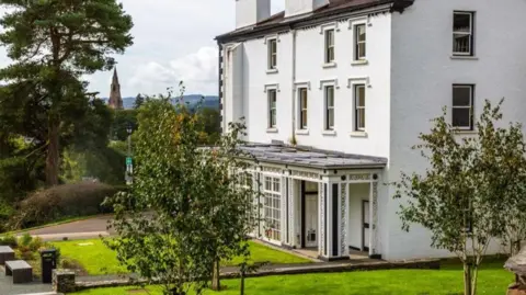 University of Cumbria Partial side and front view of a large white three storied building, dating from the 1890s. A glassed in porch stretches along the ground floor, with a pillar next to the entrance, and there are four sash windows on the front of the first storey and three on the second, and one to the side of each. An area of lawn is to the front with a large pine tree, and two smaller ones. In the distance a church tower can be seen, with hills in the far distance.