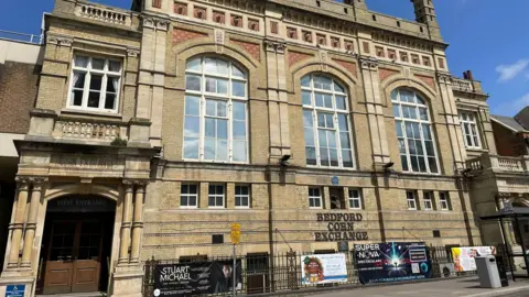 A view of an old brick building with huge windows and the words "Bedford Corn Exchange" on the side under a blue plaque which is too small to read.