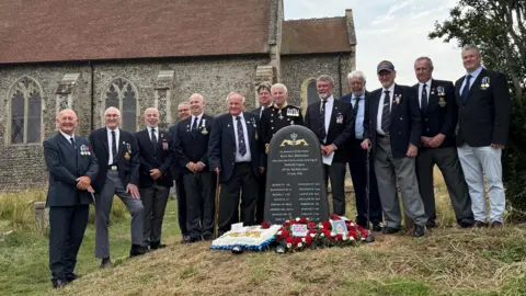 Submariners Association Men dressed smartly in navy blazers, with medals, line up behind a new gravestone-shaped memorial, with Mundesley church behind them