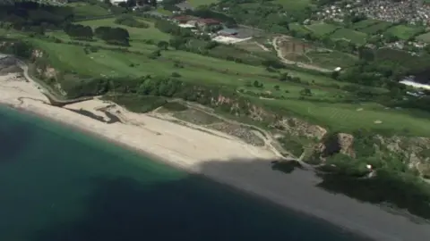 An aerial view of a scenic coastal area which features a sandy beach bordered by clear blue-green water, cliffs and green fields. A residential area is visible in the upper right corner of the image.