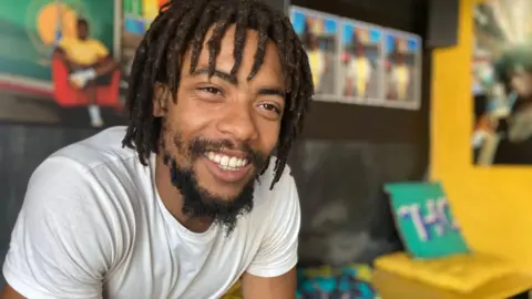 Donovan Charles Lansiquot sits in a community centre, smiling at the camera. There is bright artwork behind.