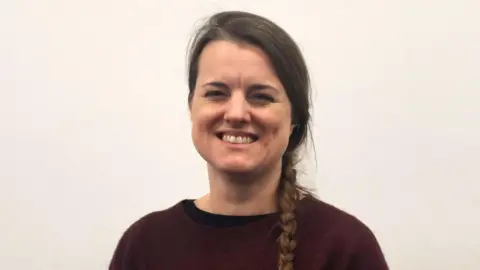Dr Sally Gillespie is wearing a maroon jumper with grey cuffs. She is wearing dark trousers and has brown hair, styled in a single plait over one shoulder. She is standing in front of a white wall.