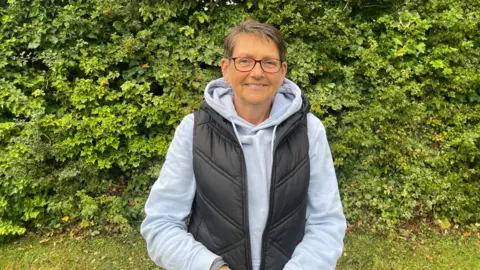 Holmes Chapel Pride committee member Alison Wright, wearing a grey hoodie and black gilet, smiles as she poses for the camera.