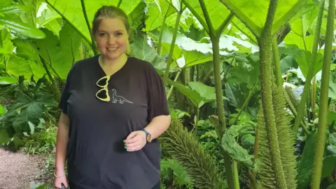 West Suffolk NHS Foundation Trust Ceiridwen is standing in a garden with lots of tall green plants behind her. She has her blonde hair tied back. She is wearing a black T-shirt and has a pair of yellow heart-shaped sunglasses tucked into her collar. 