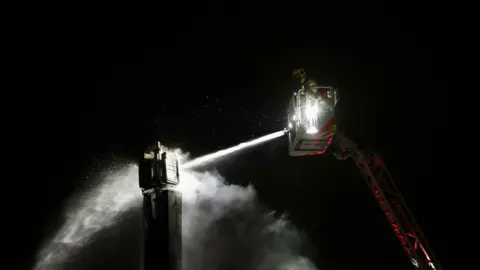 Pacemaker Press A firefighter is in an extension apparatus spraying liquid on the top of a burned 5G mast