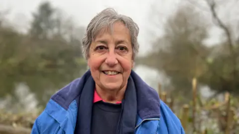 Pat Farrow, wearing a blue winter coat, blue scarf and jumper looks into the camera while in front of the lake.